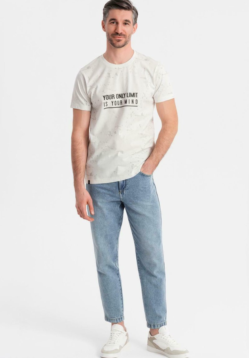 Camiseta blanca con un patrón texturizado, que presenta un texto impreso en negro: "TU ÚNICO LÍMITE ES TU MENTE." Combinada con jeans de color azul claro y zapatillas blancas.