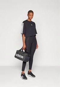 Γυναίκα σε μαύρη αθλητική ένδυση Adidas και αθλητικά παπούτσια, κρατώντας μια μαύρη τσάντα duffel Adidas, στο χέρι, μπροστά σε λευκό φόντο.