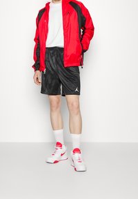 Veste coupe-vent rouge et noire, t-shirt blanc, short noir avec un motif graphique, et chaussures de sport blanches et rouges avec des chaussettes montantes.