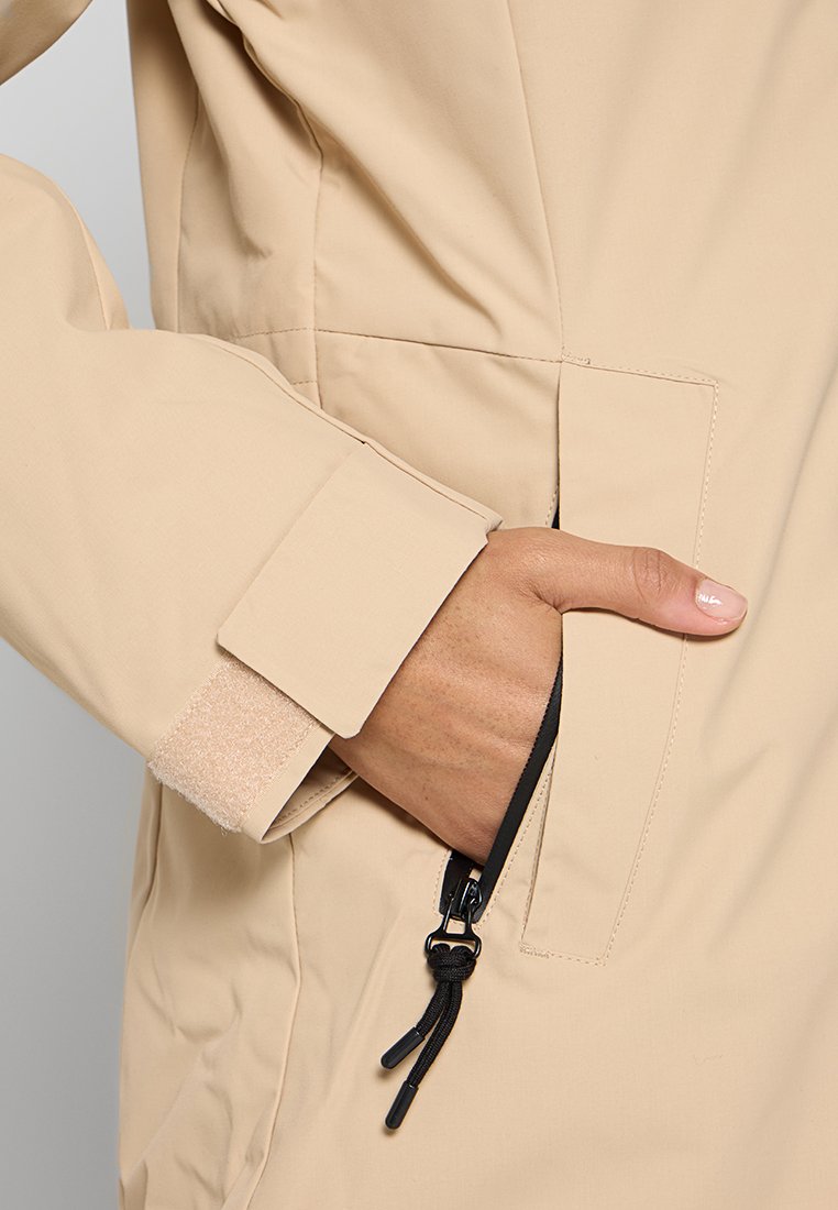 Beiger Blazer aus einem glatten Stoff, ausgestattet mit einer Reißverschlusstasche und einem mit Klettverschluss verstellbaren Ärmelbündchen. Detaillierte Nähte sind in der Nähe der Säume sichtbar.