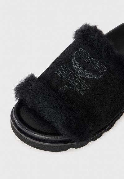 Sandal cu talpă definitivă căptușită cu blană neagră, având un superior din suede moale, cu un suport pentru picior texturat și o talpă groasă din cauciuc negru. Cusătura decorativă este vizibilă.