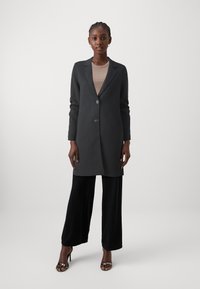Manteau gris foncé avec un col à revers, fermeture à un bouton, et deux poches latérales, associé à un pantalon large noir et des talons avec lanières.
