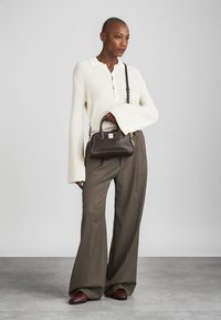 Sac à main en cuir marron avec double poignée et accents en métal doré. Le modèle porte un t-shirt à côtes blanc et un pantalon large gris.