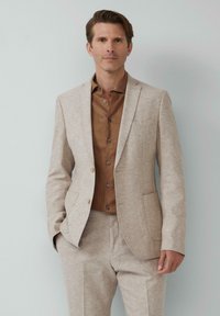 Beige colbert met textuurstof, enkele knoopsluiting en notch-lapels, gecombineerd met een bruine overhemd en bijpassende broek.