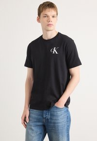 Black cotton crew-neck t-shirt featuring a white "cK" logo on the left chest; paired with light blue denim jeans.