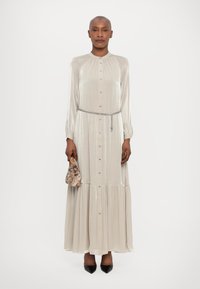 Robe longue argentée métallique avec une série de boutons, un col froncé et une taille élastique, associée à un sac à main à motifs et des chaussures noires.