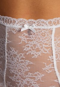 Lingerie en dentelle blanche à motifs floraux, avec un bord festonné et un petit nœud en satin à la taille. Texture délicate et transparente.