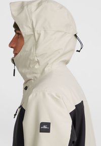 Veste légère et imperméable en crème et noir, avec capuche, fermeture éclair et patch logo sur l'épaule. Texture lisse.