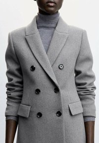 Grå uld dobbeltbrystet frakke med sorte knapper, skåret revers og sidelommer; båret over en grå turtleneck. Glat tekstur.