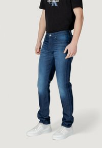 Jeans de mezclilla azul oscuro con corte ajustado, que cuentan con un cierre de botón, bolsillos frontales y un ligero desgaste; combinados con zapatillas blancas y una camiseta negra.