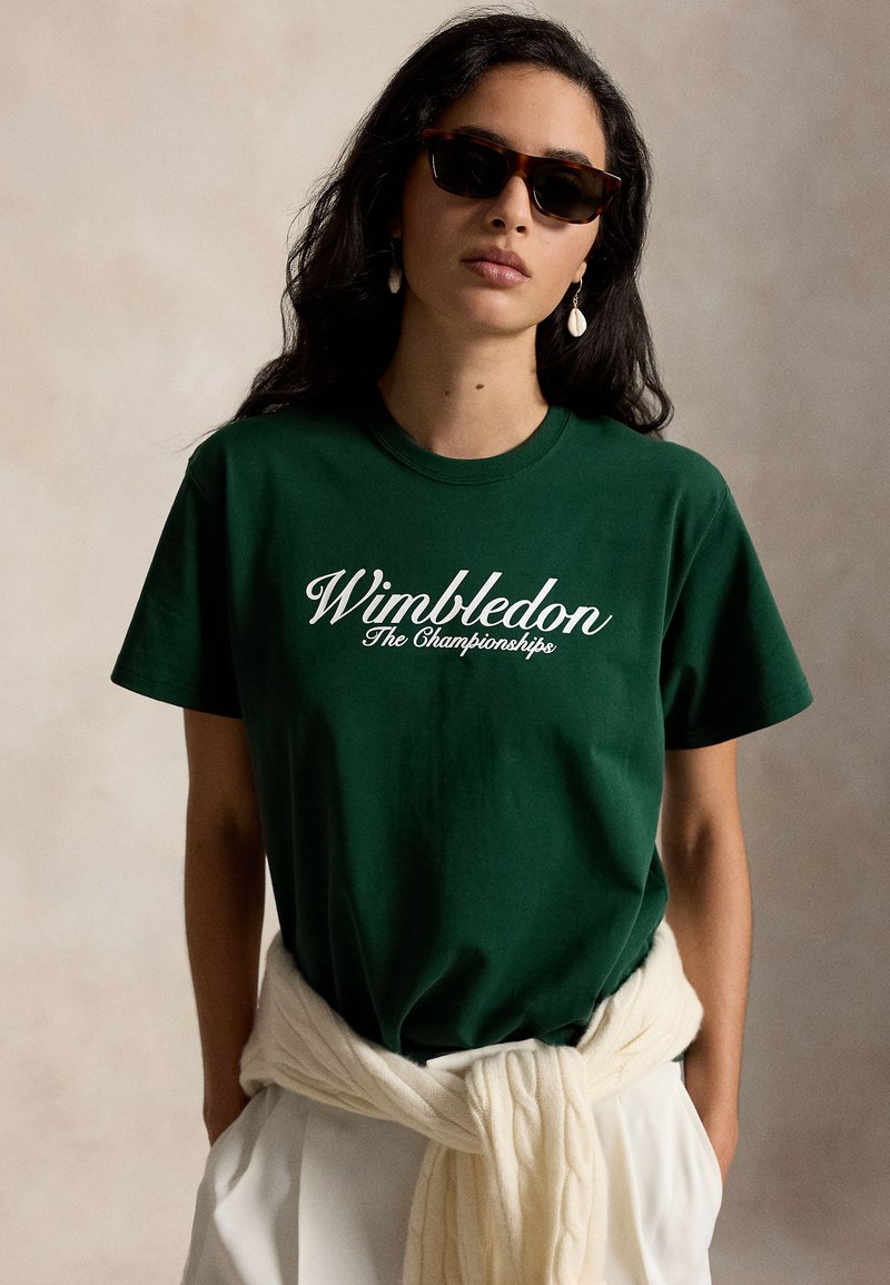 Γυναίκα με σκούρο πράσινο μπλουζάκι «Wimbledon The Championships», σκουρόχρωμα γυαλιά ηλίου, σκουλαρίκια σε σχήμα κοχυλιού και κρεμασμένο γύρω από τη μέση κρεμ πουλόβερ.