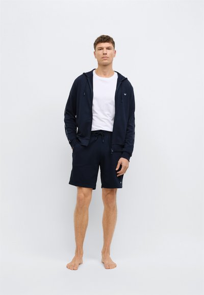 Tengerészkék cipzáras kapucnis pulóver húzózsinórral, fehér póló fölött viselve, hozzáillő tengerészkék rövidnadrággal párosítva; letisztult dizájn, látható mintázat nélkül.