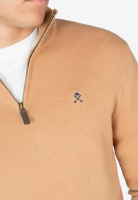 Suéter de cremallera en tono marrón claro con un acabado texturizado, que presenta un logo azul bordado en el pecho y un tirador de cremallera en color marrón.
