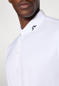 Hvit polo skjorte med strukturert krage, tre knapper og en svart logo brodert på kragen. Glatt stofftekstur.