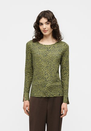 Femme aux cheveux bruns bouclés portant un haut vert à manches longues avec des taches de léopard noires et un pantalon marron, debout sur un fond uni.