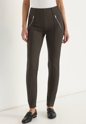 Vrouw draagt hoog getailleerde, donkerbruine slim-fit broek met verticale naden en voorzakken met rits, gecombineerd met zwarte loafers.