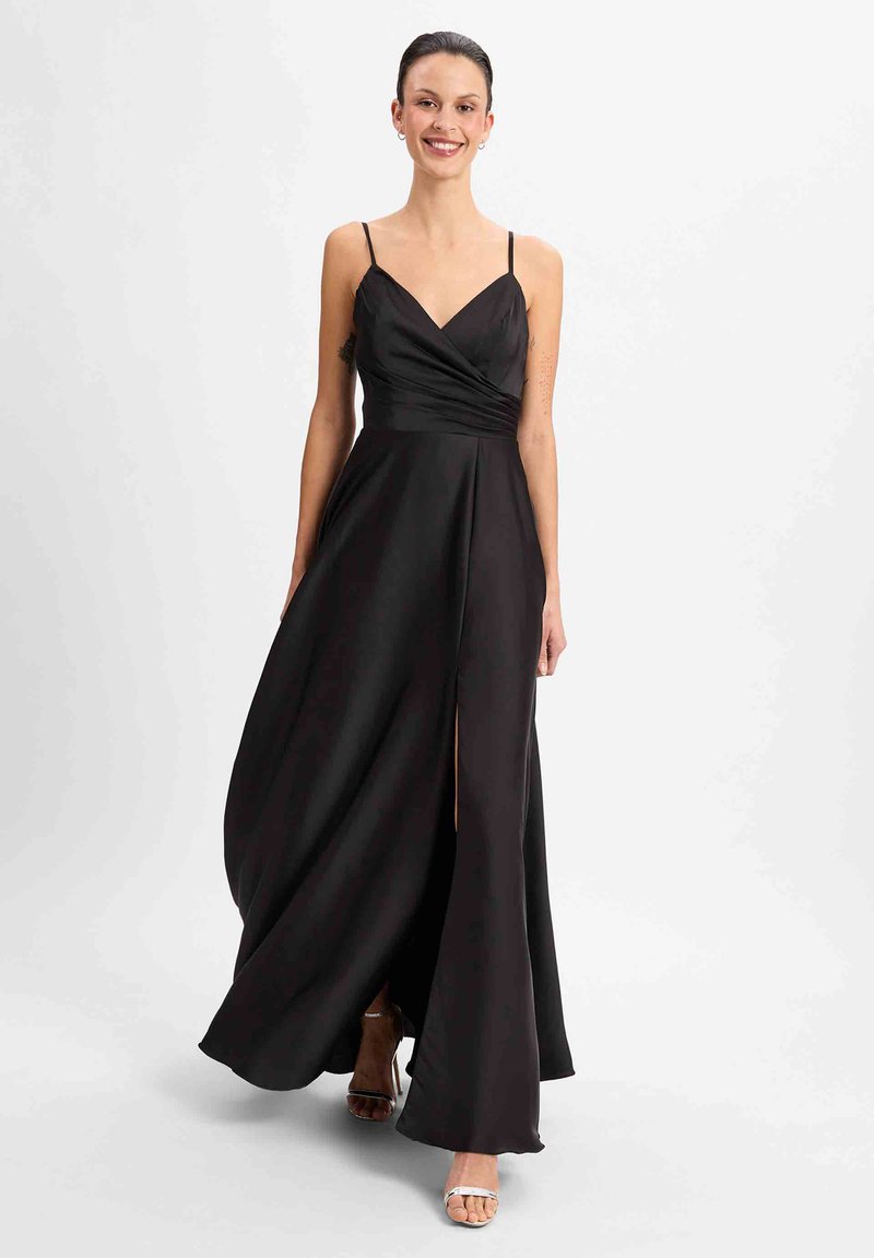 Schwarzes Satin-Kleid mit V-Ausschnitt, dünnen Trägern und einem seitlichen Schlitz. Der fließende Rockstoff ergänzt das schlichte Design. Sichtbare silberne Riemchen-Highheels.