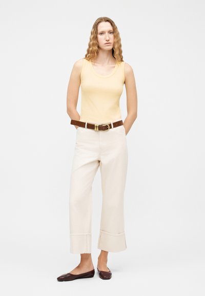 Γυναίκα με κυματιστά μαλλιά, που φοράει κίτρινο αμάνικο τοπ, εκρού παντελόνι ψηλόμεσο με ρεβέρ, καφέ ζώνη και σκούρα καφέ μπαλαρίνες, στέκεται με τα χέρια πίσω.