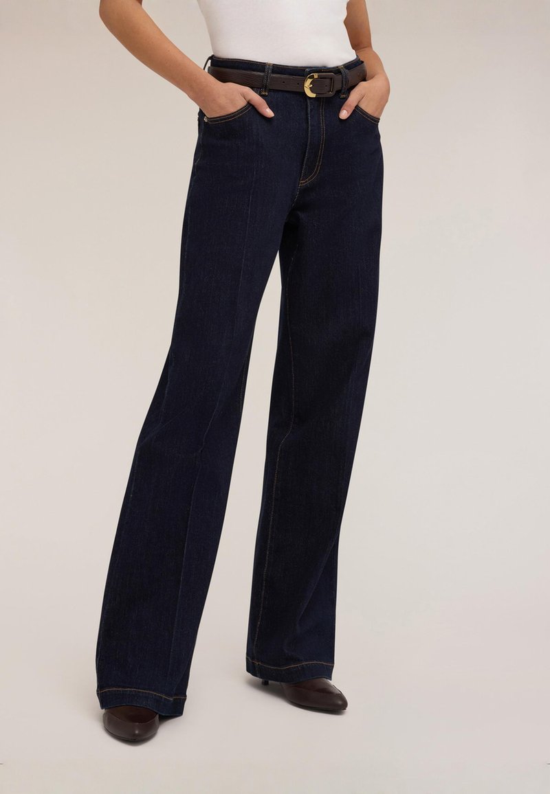 Mörkblå vida jeans med hög midja, med orange sömmar och sidofickor, kombinerade med ett brunt bälte och klackskor.