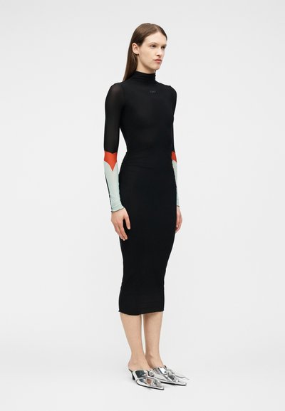 Nő áll, testhez simuló fekete ruhát visel, magas nyakkal, többszínű hosszú ujjakkal és hegyes ezüst mules cipővel.