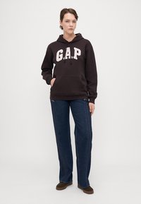 Hanorac negru cu logo "GAP" în roz deschis, buzunar frontal, glugă cu șnur; asortat cu blugi largi de culoare închisă și pantofi maronii.