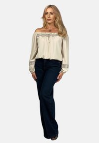 Bluzka w kolorze kremowym z odkrytymi ramionami i koronkowymi akcentami; zestawiona z ciemnoniebieskimi dżinsami typu dzwony. Gładka tekstura, luźny krój i szczegółowe mankiety.