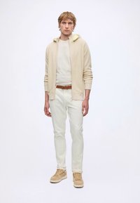 Jeune homme portant un sweat à capuche beige zippé, un t-shirt blanc, un pantalon blanc, une ceinture marron et des baskets beiges, debout sur un fond blanc uni.