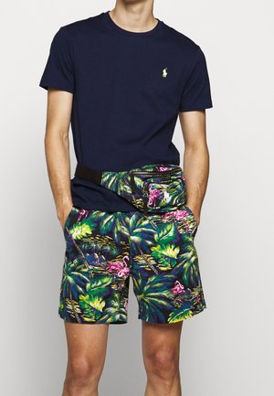 Homme portant un t-shirt bleu marine avec petit logo et un short à imprimé tropical avec des flamants roses, assorti à une sacoche de taille.