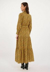 Robe maxi florale jaune avec des motifs verts et rouges, manches longues, jupe à volants, taille cintrée, portée avec des bottines noires.