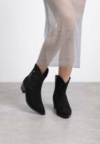 Las botas negras de ante hasta el tobillo presentan una punta afilada y acentos decorativos texturizados en negro, con un ligero tacón. Una gruesa tela transparente cae alrededor de las piernas.