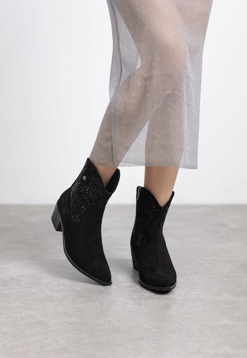 Las botas negras de ante hasta el tobillo presentan una punta afilada y acentos decorativos texturizados en negro, con un ligero tacón. Una gruesa tela transparente cae alrededor de las piernas.