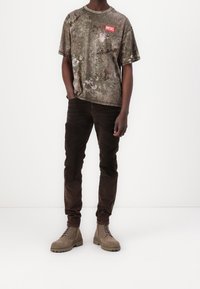 Mann trägt ein T-Shirt mit Camouflage-Muster von Diesel, dunkle braune Hose und beigefarbene Schnürstiefel und steht vor einem schlichten weißen Hintergrund.