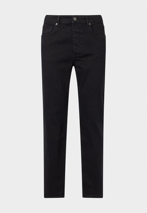 Homme portant une chemise noire à boutons, un t-shirt blanc, un pantalon noir et des baskets noires, debout devant un fond clair uni.