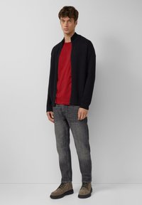 Donker marineblauw vest met ritssluiting over een rode T-shirt, gecombineerd met grijze spijkerbroek en lichtbruine veterschoenen. Zachte texturen en moderne pasvorm.