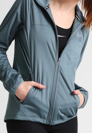 Personne portant une veste bleu à fermeture éclair sur un haut noir avec un logo blanc, les mains dans les poches de la veste.