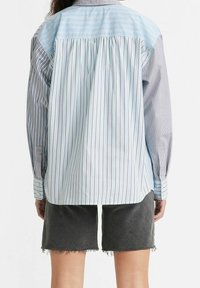 Camicia a righe con pannelli blu e grigi, caratterizzata da un design con pieghe sulla schiena e maniche lunghe, abbinata a pantaloni corte grigi scuri.