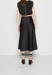 Femme portant un crop top noir sans manches avec une fermeture éclair dorée et une jupe midi en satin noire, assortis à des sandales à talons carrés noires avec des lacets.