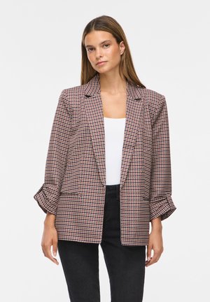 Kvinde med langt brunt hår iført en mønstret blazer over en hvid top og sorte bukser, står mod en ensfarvet lys baggrund.