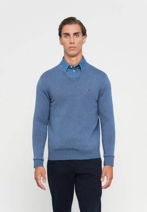Homme portant un pull bleu en V sur une chemise en jean et un pantalon sombre, se tenant devant un fond clair uniforme.
