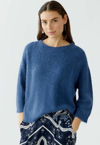 Pull en tricot bleu avec une texture douce, un col rond et des manches trois-quarts, associé à un pantalon sombre à motifs.