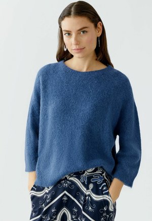 Blauer Strickpullover mit weichem Material, rundem Ausschnitt und Dreiviertelärmeln, kombiniert mit gemusterten dunklen Hosen.