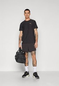 Zwart gestreept T-shirt met korte mouwen, donkere korte broek en zwarte sportschoenen. Draagt een zwarte duffeltas, met zichtbare tatoeages.