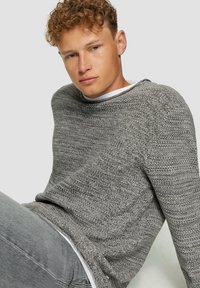 Grijze gebreide trui met een brede halslijn, tekstuurpatroon en lange mouwen. Draagt over een witte shirt met grijze jeans.