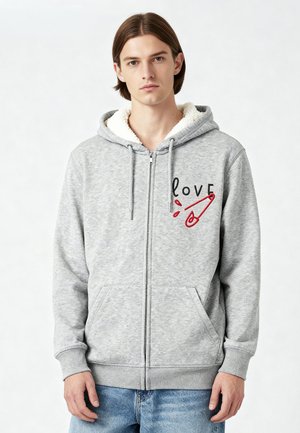 Grijze zip-up hoodie met een gevoerde capuchon, voorzakken en een grafisch ontwerp met "love" en een veiligheidsspeld in het rood. Zachte textuur.