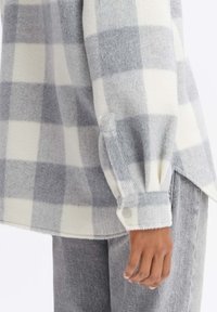 Camisa de franela a cuadros en gris y blanco con una textura suave, puños abotonados y dobladillo redondeado, combinada con pantalones grises.