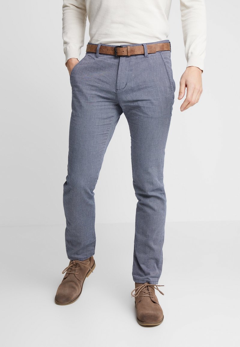 Homme portant un pantalon bleu clair texturé, une ceinture marron, des chaussures à lacets marron clair et un haut blanc à manches longues, une main dans la poche.