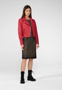Veste de moto en cuir rouge avec des détails zippés, superposée à une robe en dentelle noire. Le modèle porte des bottes noires à lacets. Arrière-plan simple.