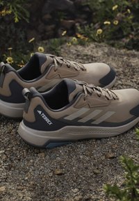 Brązowe i szare buty trekkingowe z texturedowym materiałem wierzchnim, amortyzowaną podeszwą oraz wyraźnym logo Adidas. Posiadają wzmocniony zapiętek i system sznurowania.