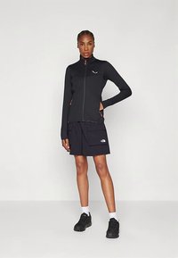 Juoda lengva striukė su užtrauktuku, aukštu apykakliu, briaunotu audiniu ir oranžiniais akcentais. Derinama su tamsiomis šortais ir juodais sportiniais bateliais.