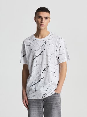 Giovane con capelli corti che indossa una maglietta bianca con crepe nere simili al marmo e jeans grigi, in piedi davanti a uno sfondo bianco uniforme.